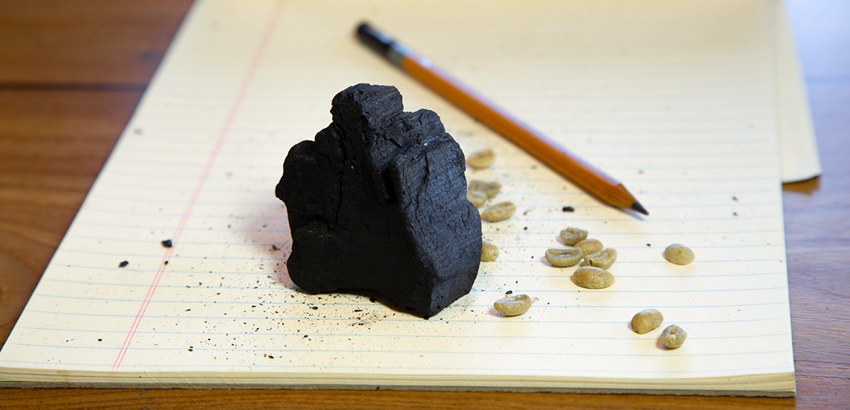 Feature image of a piece of charcoal and a graphite pencil representing carbon, alongside green coffee beans.